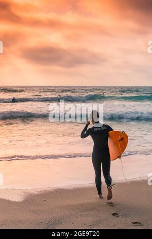 A female surfer carrying her surf board and walking into the sea in late evening sunlight at Fistral Beach in Newquay in Cornwall. Stock Photo