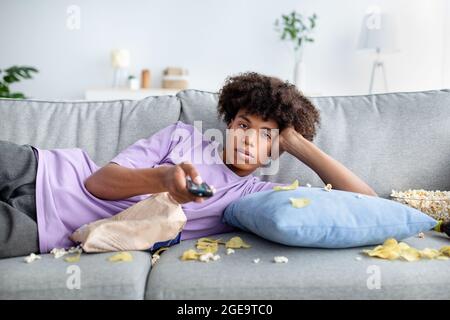 Lazy black teen guy lying on couch with scattered snacks, holding remote control, watching TV at home Stock Photo