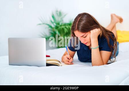 Young female student lying on bed and writing in copybook while studying online with laptop during distance education at home Stock Photo