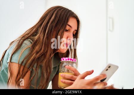 Side view of young female student browsing social networks on mobile phone near table with fresh fruits and juice while spending morning at home Stock Photo