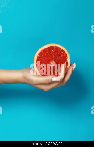 Crop anonymous female with manicure holding half of fresh juicy grapefruit with red pulp against blue background Stock Photo