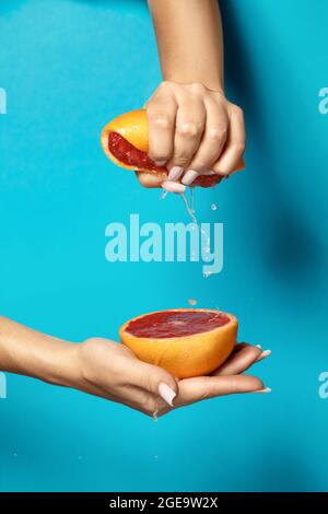 Crop anonymous female with manicure squeezing half of fresh ripe juicy grapefruit against blue background Stock Photo