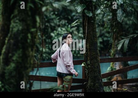 Back view of young female explorer looking over shoulder with interest while standing on wooden footbridge over Celeste river with turquoise water sur Stock Photo