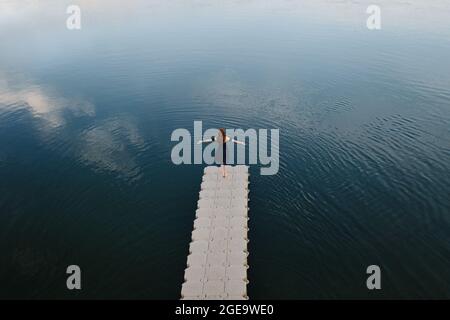 High angle back view of female standing with outstretched arms on quay near lake and enjoying freedom Stock Photo