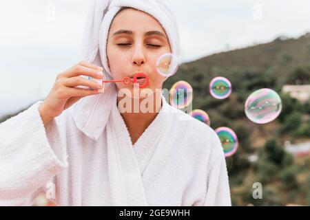Young Woman in white bathrobe and towel blowing soap bubbles during spa session on balcony Stock Photo