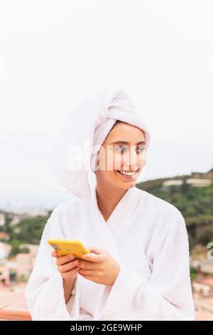 Young female in bathrobe and towel smiling and browsing mobile phone while resting on balcony after shower Stock Photo