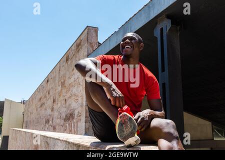 Low angle of smiling African American male athlete in activewear sitting with bottle of water on stone border in street during training and looking aw Stock Photo