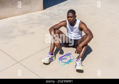 High angle of tired African American male athlete sitting with jumping rope on sports ground and taking break during workout in summer Stock Photo