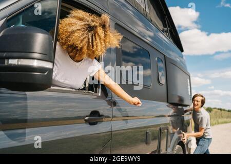 Young man pushing camper van while Woman sitting at steering wheel as having troubles during trip in summer countryside Stock Photo
