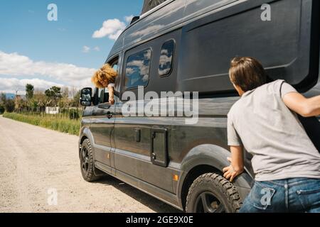 Back view of young man pushing camper van while Woman sitting at steering wheel as having troubles during trip in summer countryside Stock Photo