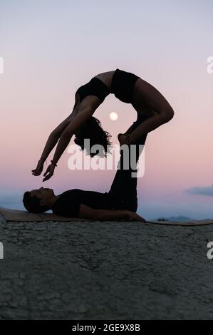 Side view of silhouette of flexible Woman doing backbend and balancing on legs of man during acroyoga session against sunset sky with moon Stock Photo