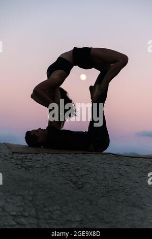 Side view of silhouette of flexible Woman doing backbend and balancing on legs of man during acroyoga session against sunset sky with moon Stock Photo