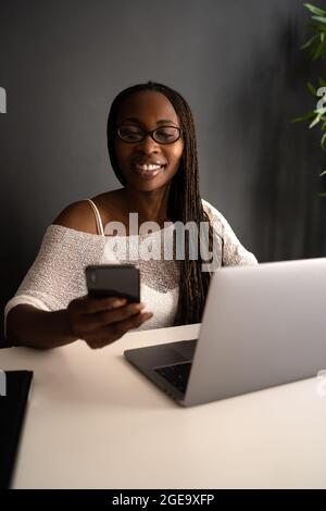 Cheerful African American female freelancer sitting at table in modern workplace and browsing cellphone while working on project remote from home Stock Photo