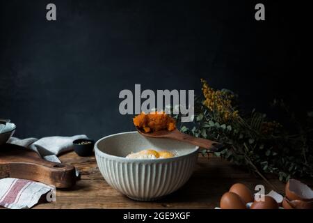Bowl with eggs, flour and wooden spoon with pumpkin puree on wooden table during pastry preparation on dark background Stock Photo