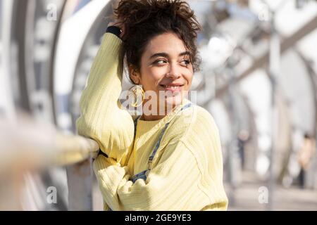 Confident Hispanic female teenager with curly hair wearing denim overalls and yellow sweatshirt with earrings looking away while leaning on railing on Stock Photo