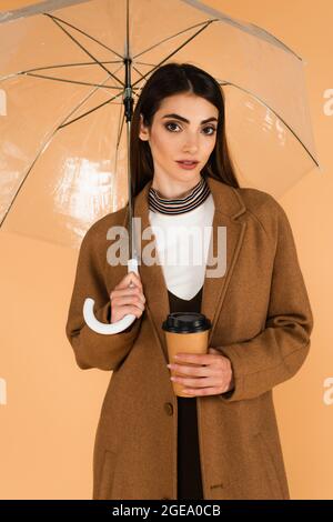 young woman in brown coat standing with paper cup under transparent umbrella isolated on beige Stock Photo