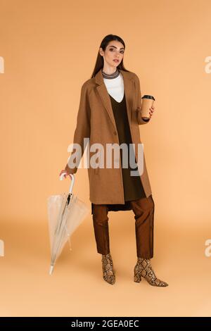 brunette woman in fashionable autumn clothes posing with paper cup and umbrella on beige Stock Photo
