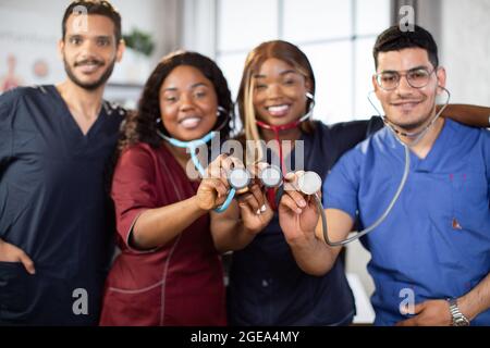 Blur front view of four young diverse multiethnic medical practitioners, standing together in light hospital room and demonstrating stethoscopes to camera. Focue on stethoscope heads Stock Photo