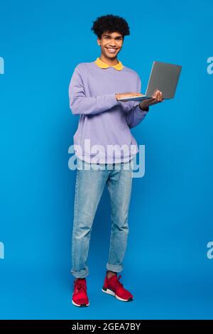 full length view of african american student using laptop on blue background Stock Photo