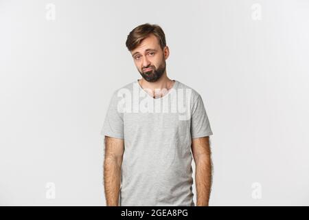 Portrait of sad and gloomy bearded man in gray t-shirt, frowning and looking miserable, standing over white background Stock Photo