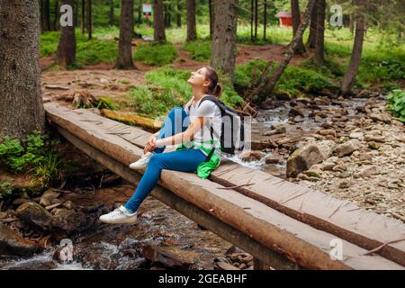 Traveler hiker with backpack crossing mountain river in Carpathian forest walking on wooden bridge. Woman tourist enjoying natural landscape. Trip to Stock Photo