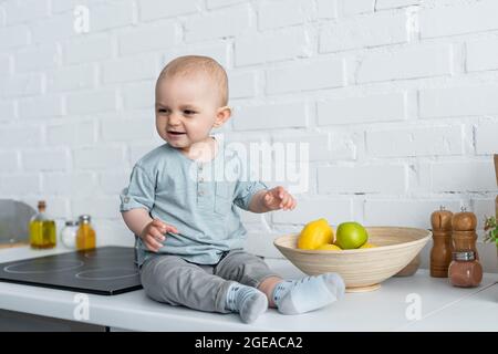 Smiling baby sitting near fruits on kitchen worktop Stock Photo