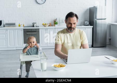 Man using laptop near breakfast and baby on high chair in kitchen Stock Photo