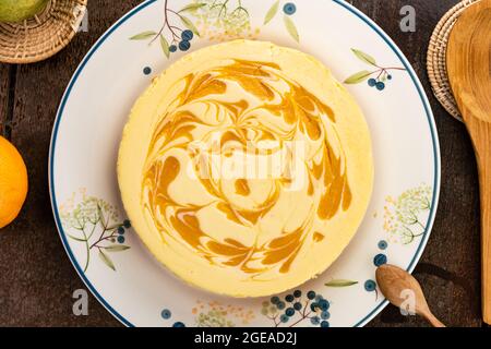 Top view of delicious homemade mango cheesecake in ceramic dish with mango, ripe orange, wooden spoon, bamboo tray on wooden table. Stock Photo