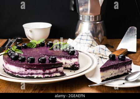 Piece and the whole of delicious homemade blueberry cheesecake garnished with preserved blueberry and green mint leat in ceramic dish with metal spoon Stock Photo