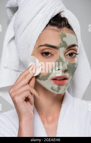 young woman in bathrobe removing clay mask with cotton pad isolated on grey Stock Photo