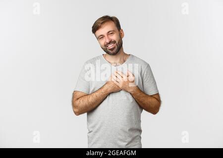 Image of handsome kind man with beard, wearing gray t-shirt, holding hands on heart and sighing, feeling thankful, express gratitude, standing over Stock Photo