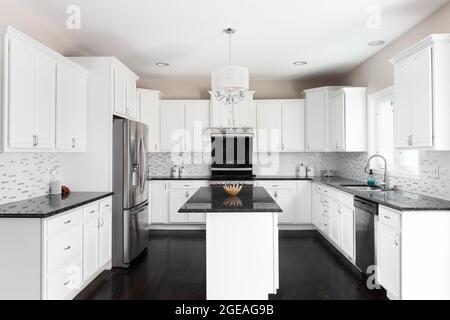 A large white kitchen with dark bamboo floors, black granite countertops, a light hanging above the island, and stainless steel appliances. Stock Photo