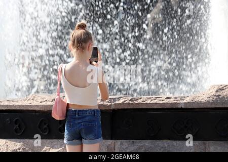 Kid girl photographs on smartphone camera water jets of fountain. Child in summer, hot weather in city Stock Photo
