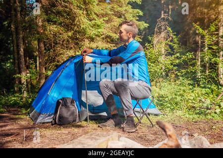Young man setting up tent on camping trip in forest. Summer camping with campfire. Traveler adjusting tent elements. Stock Photo