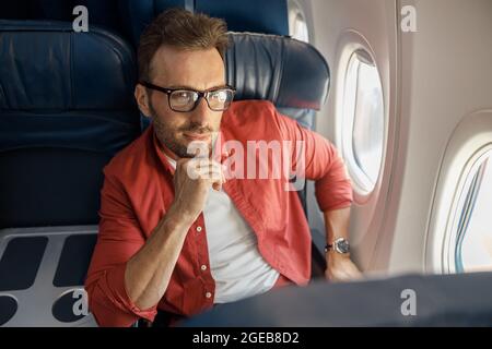 Attractive caucasian businessman in casual wear and glasses looking thoughtful, sitting on the plane near the window Stock Photo