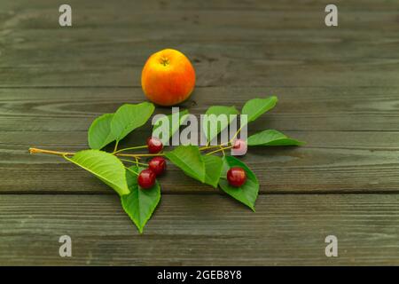 Apples placed on a black wooden substrate. Next to it, on a twig with leaves, there are cherries. Stock Photo