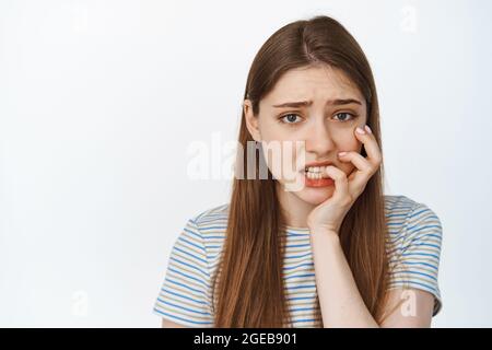 Portrait of scared girl biting her finger, looking anxious and worried at camera, standing in t-shirt against white background Stock Photo