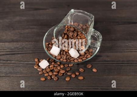 A glass cup filled with brown coffee beans. It stands on a dark surface with the saucer. There are two white sugar cubes on top of the coffee beans. Stock Photo