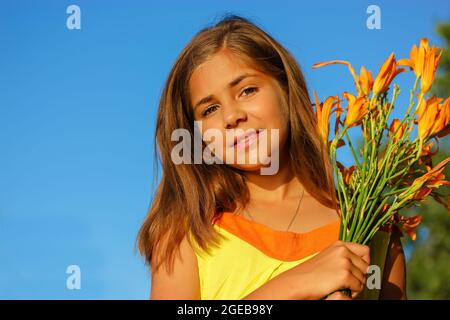 A beautiful happy teenage girl in yellow dress with a bouquet of orange lilies against a blue sky at sunny summer day. Female kid of 10-12 years old. Stock Photo