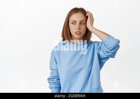 Image of young confused blond girl trying to understand smth, scratch head and look clueless aside, thinking, making up plan, standing against white Stock Photo