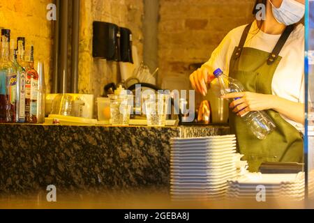 A bistro cafe staff, female waitress, wearing a protective medical mask prepares a takeaway cocktail. An unrecognizable barista in coffee shop. Corona Stock Photo