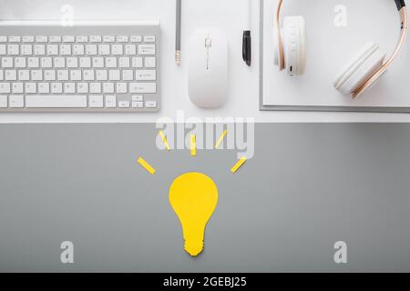 Yellow light bulb idea concept on desktop with keyboard pc computer mouse headphones and white suppliers in workspace on gray table background. Stock Photo
