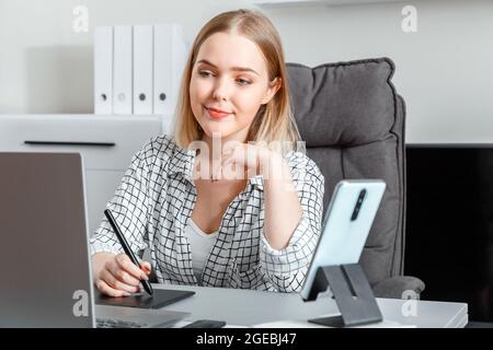 Confident young woman designer using graphic tablet laptop smartphone for work in home office workspace interior. Freelance retoucher illustrator Stock Photo