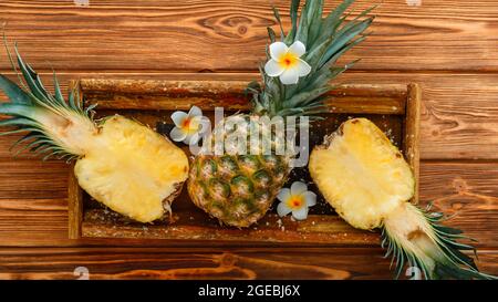 Ripe Pineapple. Tropical summer fruit pineapple halves and whole pineapple on brown dark table in wooden box with tropical plumeria flowers. Long web Stock Photo