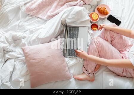 Young woman in pajamas sitting in bed using laptop for remote work and smartphone while drinking tea having breakfast. Woman at home workspace in bed Stock Photo