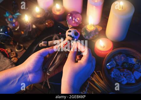 Sorceress or witch sticks needles into voodoo doll at ritual table with pentagram, burning candles and other occult objects, top view. Voodoo witchcraft, spirituality and occultism concept. Stock Photo