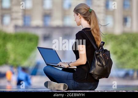 Slender woman in her 30s does remote work on street using laptop. Stock Photo