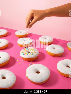 A colorful photo of a pattern of white-glazed donuts with a beautiful hand pouring multi-colored sprinkles over one of the donuts and pink background Stock Photo