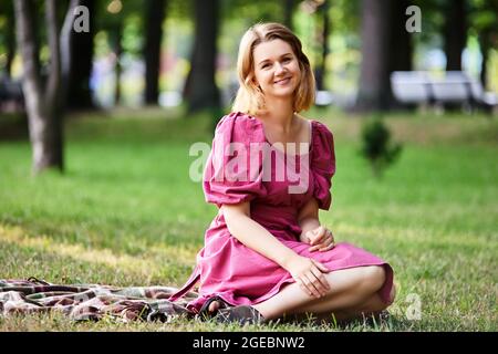 Happy woman in summer dress sits on grass in park. Stock Photo