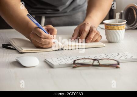 close-up of a person's hand taking notes with a blue pen on a notebook, workplace with glasses, computer mouse, and keyboard. office supplies Stock Photo